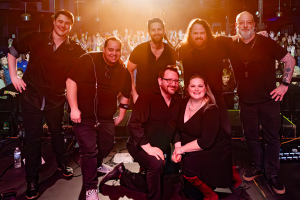 Group of seven performers posing on a concert stage after a show, with a cheering crowd and warm lighting in the background.