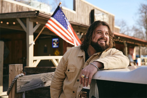 Bearded man in a beige jacket leaning on a railing beside a dock, with an American flag waving in the background.